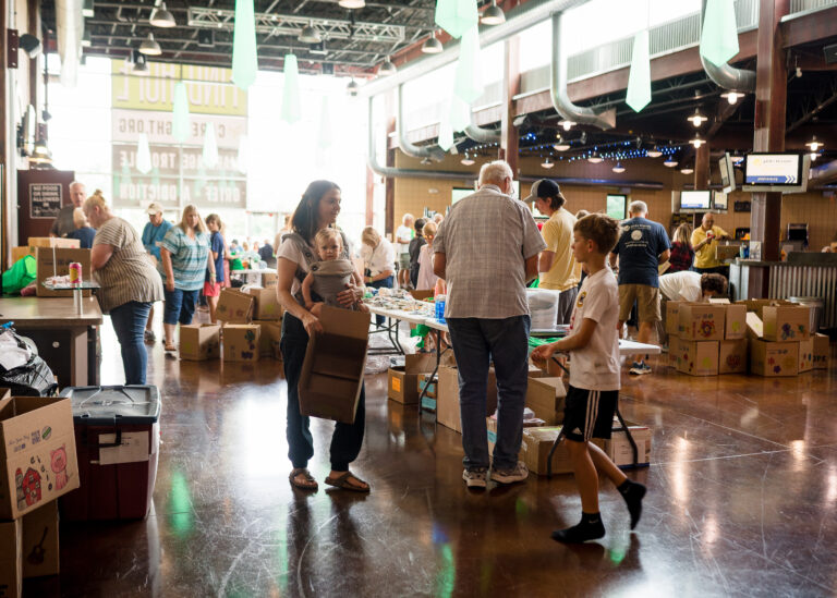 People pack boxes and organize supplies at an indoor community center, assembling Wheaton cancer care packages or sorting Chicago faith-based cancer support donations for a volunteer event.
