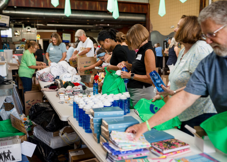 A group of people pack hygiene products, books, and supplies into bags at long tables in a community center as part of Crown Point volunteer opportunities.