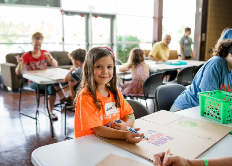 A young girl wearing an orange t-shirt and name tag sits at a table with a cardboard project in a busy classroom, highlighting Crown Point volunteer opportunities as other children and adults work at nearby tables.