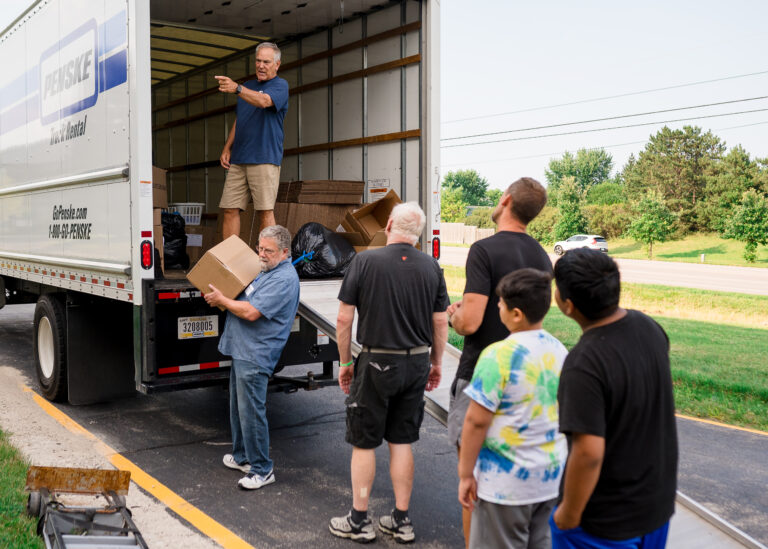 Several people stand in line by a moving truck as one man directs and another loads boxes—part of Orlando care packages—into the truck on a paved driveway near a grassy area.