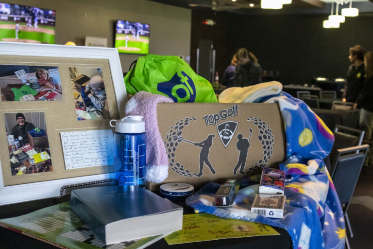 A tabletop displays a framed collage, TopGolf sign, water bottle, blankets, books, small gifts, and cards—highlighting Chicago faith-based cancer support—while TV screens in the background show a baseball game.
