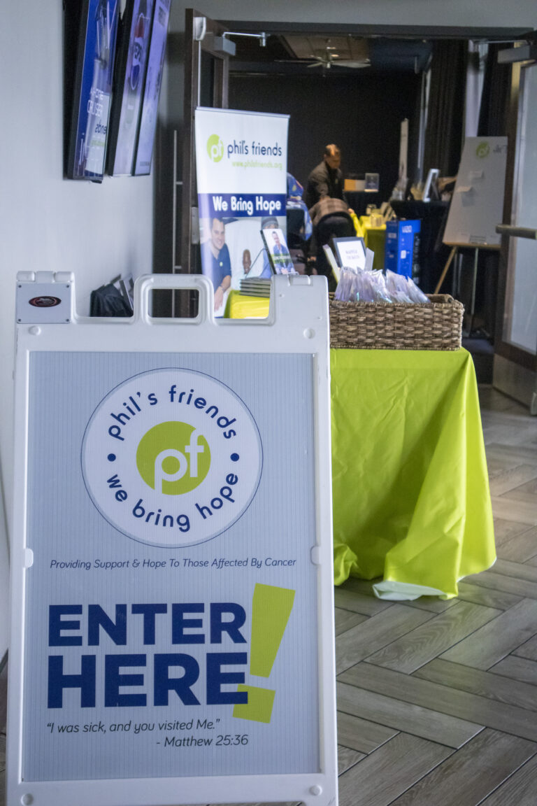 A sign reading "Phil's Friends: We Bring Hope. Enter Here!" stands in front of a registration table with informational materials about Chicago faith-based cancer support and a donation basket in a hallway.