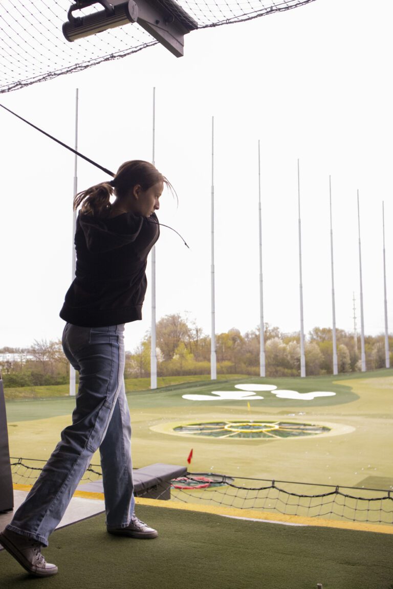 A person swings a golf club at an indoor driving range, facing an outdoor field with targets and tall netting, reminiscent of the community focus seen in Indianapolis donations.