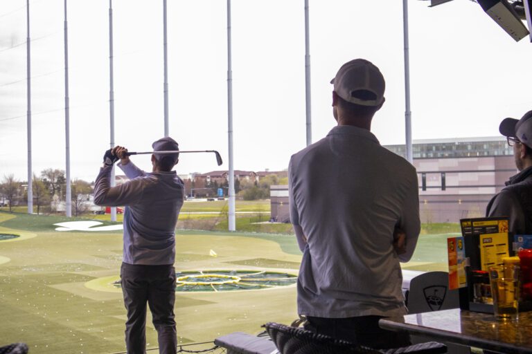 A person swings a golf club at an outdoor driving range while two others watch; tables with food and drinks, including Orlando care packages, are in the foreground.