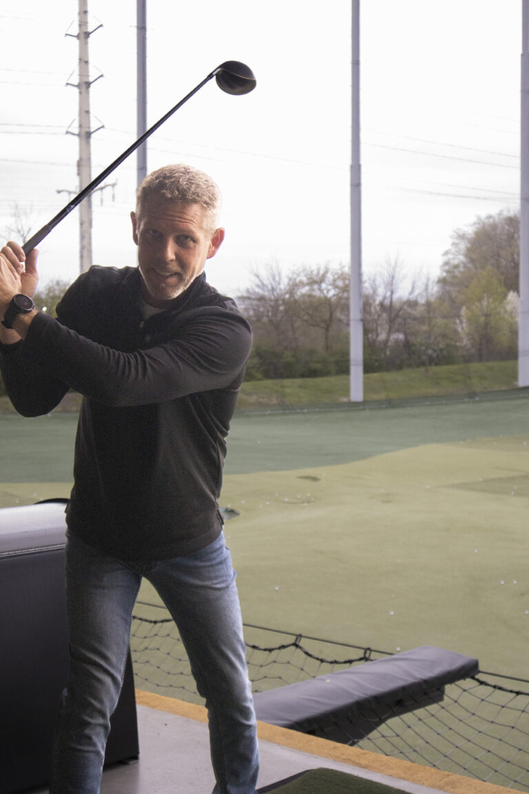 Man wearing a black shirt and jeans holding a golf club in a backswing position at a driving range, supporting Wheaton cancer care packages with every swing.