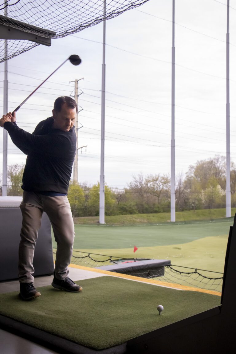 A man stands on an artificial turf mat at an outdoor driving range, preparing to swing a golf club. Nearby, Crown Point volunteer opportunities are highlighted for those interested in supporting the local community.