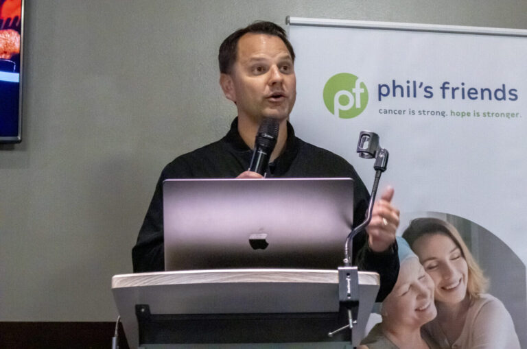 A man stands at a podium speaking into a microphone, with a laptop in front of him and a "Phil’s Friends" banner in the background, sharing about Chicago faith-based cancer support.