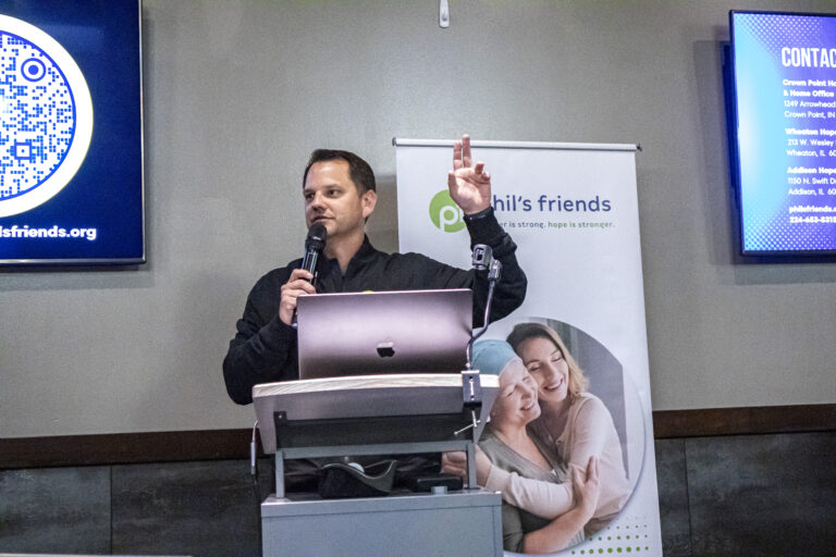 A man speaks at a podium with a microphone. Behind him are a banner showing two women hugging and a sign with contact information for Chicago faith-based cancer support.
