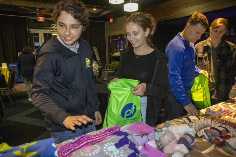 Several people stand at a table filled with assorted items, packing green drawstring bags labeled "Phil's Friends" as part of Wheaton cancer care packages at an indoor event.