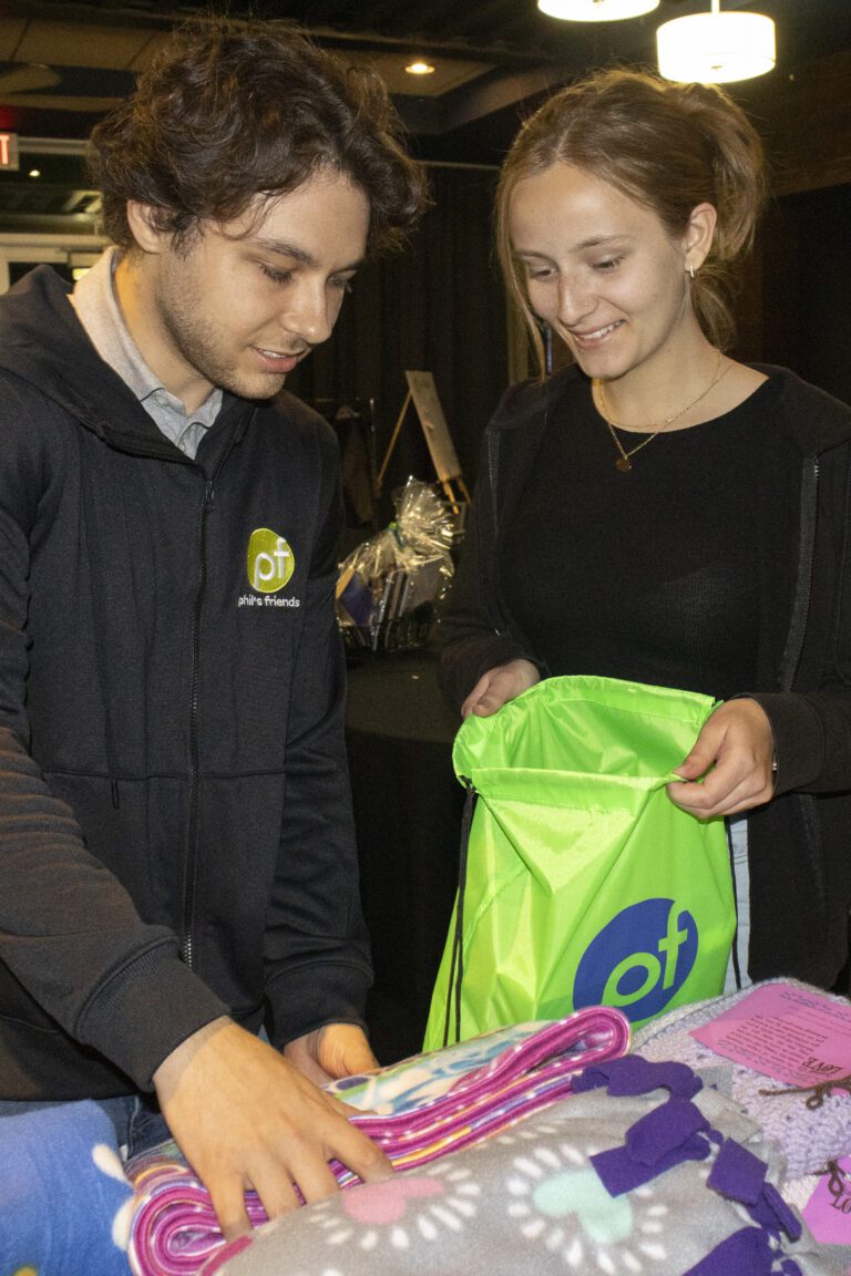 Two people stand at a table with colorful fleece blankets; one folds a blanket while the other holds a bright green drawstring bag, highlighting Crown Point volunteer opportunities.