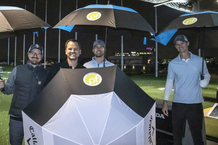 Four men stand outdoors at night under large branded umbrellas at a golf driving range, smiling at the camera while discussing Indianapolis donations.