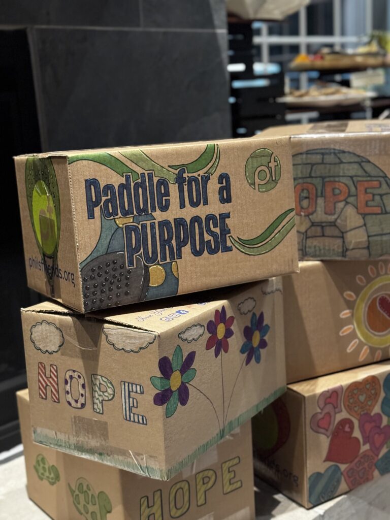 Stacked cardboard boxes decorated with colorful drawings and messages, including "Paddle for a Purpose" and "Hope," highlight Crown Point volunteer opportunities supporting local Indianapolis donations.