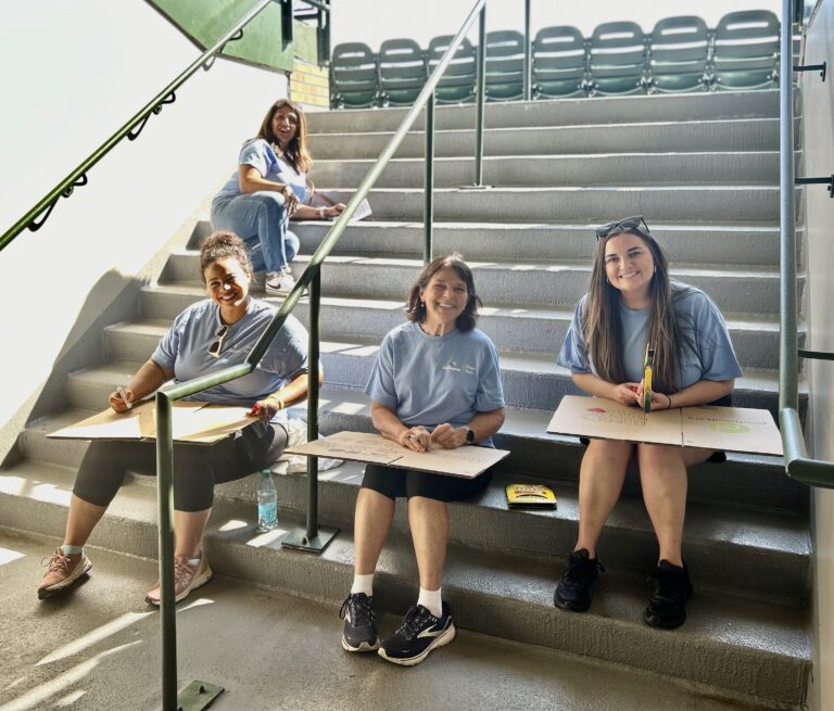 Four women in blue shirts sit on concrete steps, each holding cardboard and markers, smiling at the camera. They gather to support Wheaton volunteer opportunities. Folding chairs and a railing are visible in the background.