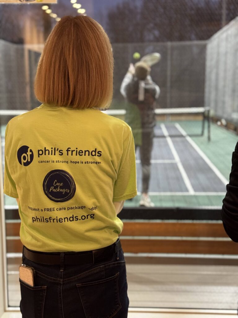 A person wearing a bright yellow "phil's friends" t-shirt watches a paddle tennis match through a glass window, highlighting Wheaton volunteer opportunities.