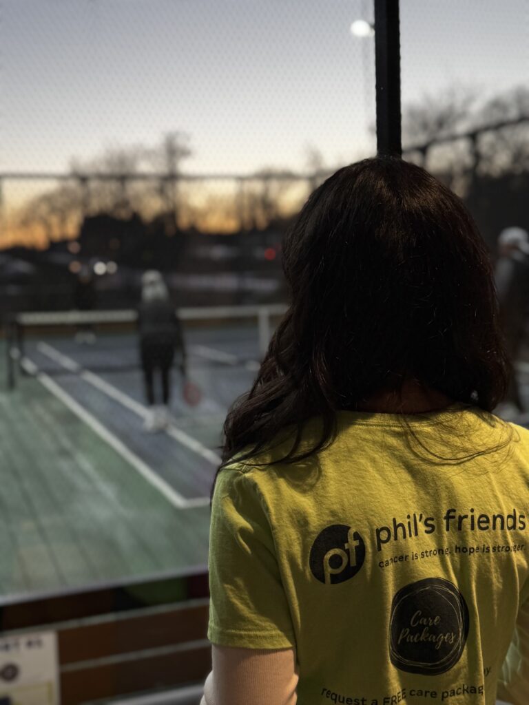 A person wearing a lime green "phil's friends" t-shirt stands indoors, looking out at people playing pickleball on an outdoor court at sunset, supporting Chicago faith-based cancer support with Wheaton volunteer opportunities.