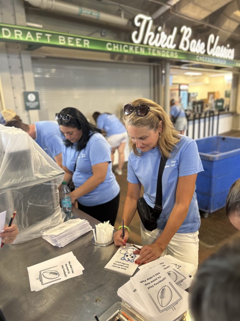 A woman in a blue shirt fills out a form at a counter with papers and pens, while other people in matching shirts—part of Crown Point volunteer opportunities—work nearby in a food court area.