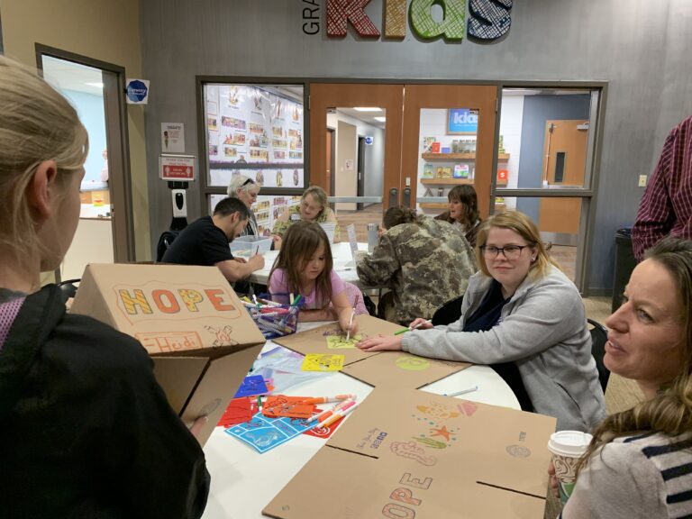 Adults and children sit at a table decorating cardboard boxes with markers and stickers in a community center or classroom, assembling Orlando care packages to support those in need.