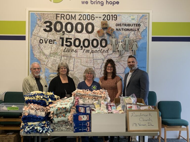 Five people stand in front of a display showing distribution statistics, with tables of donated blankets and Orlando care packages labeled "Landmark Church Mt. Vernon, IL.