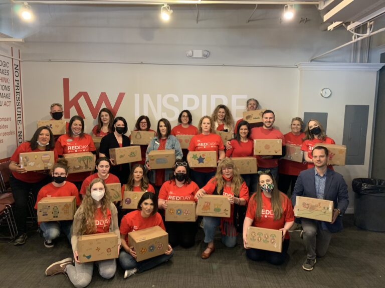 A group of people in matching red shirts pose indoors holding decorated cardboard boxes, supporting Crown Point volunteer opportunities in front of a wall with "kw INSPIRE" signage.