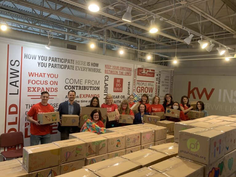 A group of people stand behind a table filled with decorated cardboard boxes in a well-lit room with motivational wall quotes—part of Wheaton cancer care packages and local volunteer opportunities.