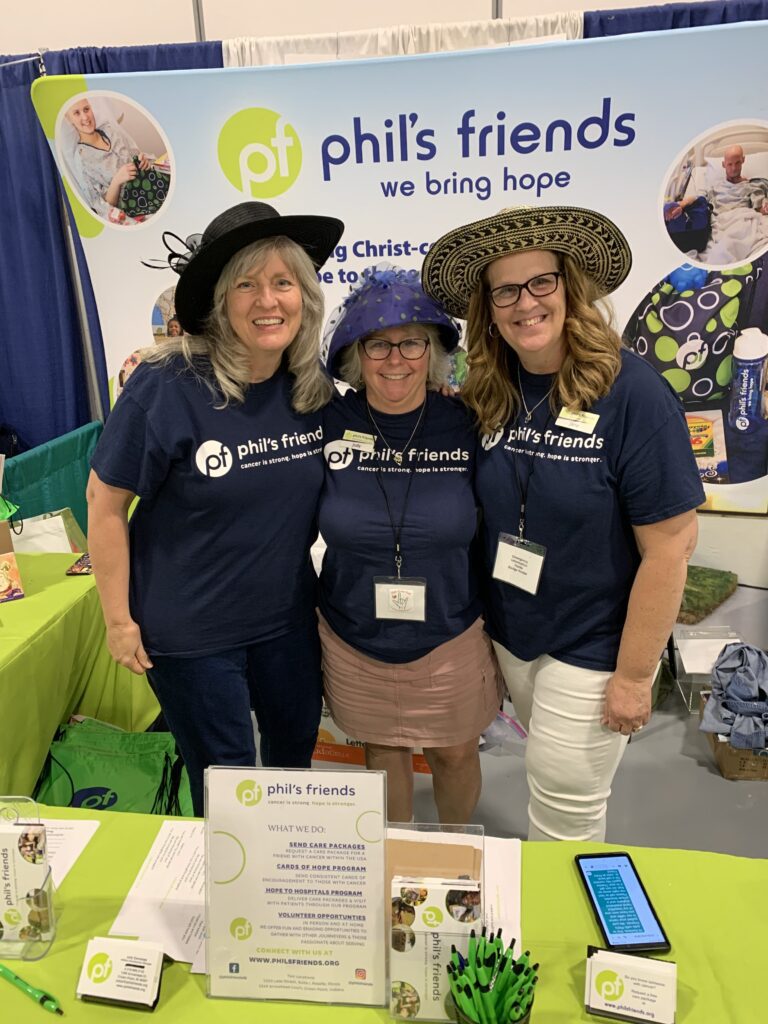 Three women wearing hats and "phil's friends" t-shirts stand and smile behind a display table at a booth promoting the organization's mission and sharing information about Crown Point volunteer opportunities.