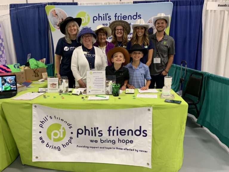 Eight people wearing hats stand behind a table covered with a green cloth promoting Phil's Friends, an organization supporting those affected by cancer through Wheaton cancer care packages.