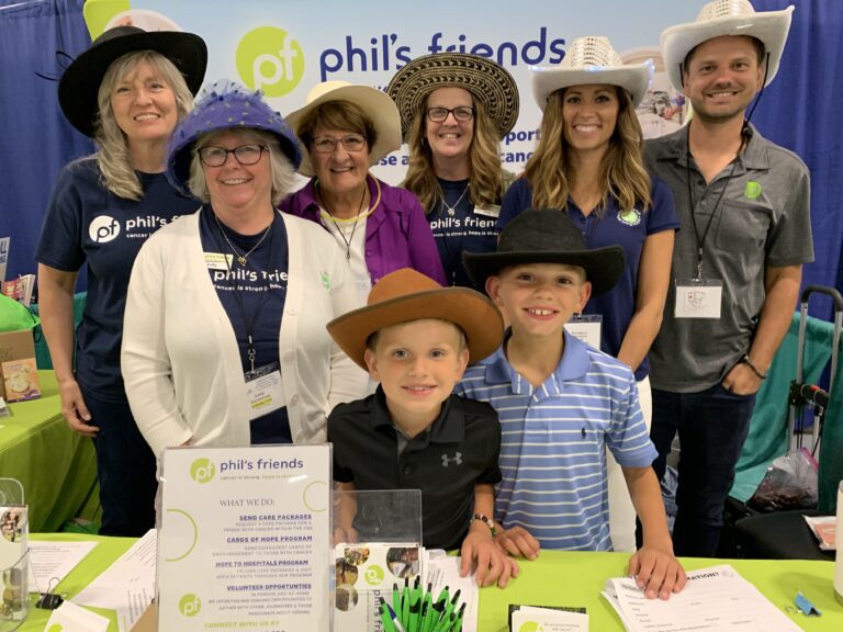 A group of adults and children wearing hats stand smiling behind a table at a Phil's Friends booth, sharing informational materials and pens about Chicago faith-based cancer support.