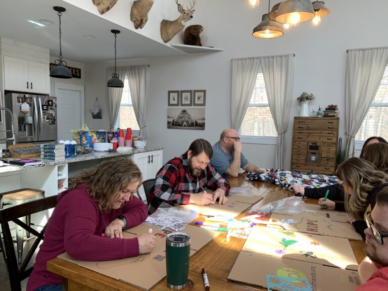 Five adults sit around a wooden table in a bright kitchen, drawing on cardboard with markers to assemble Wheaton cancer care packages. Art supplies and drinks are scattered on the table, with animal mounts displayed on the wall.