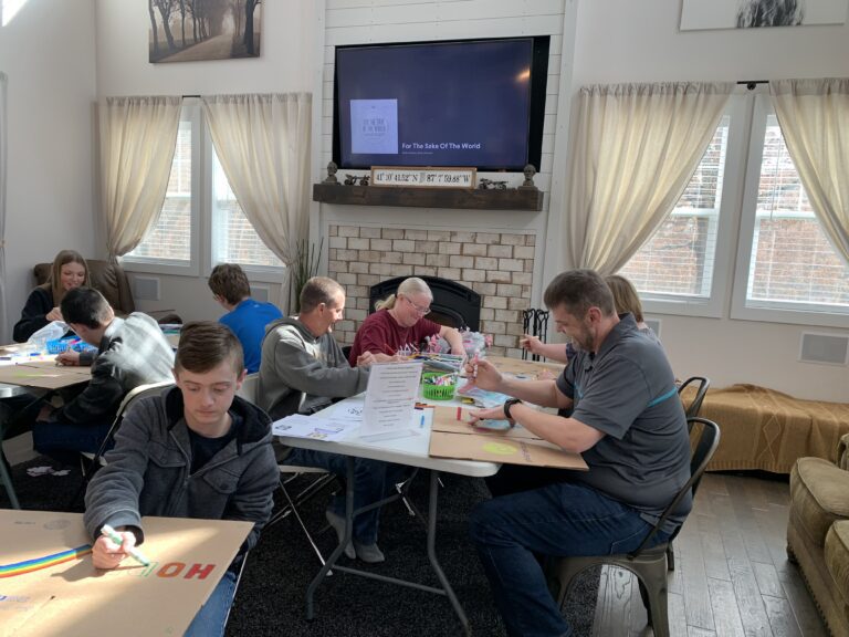Several people sit around tables in a well-lit living room, drawing and coloring on large sheets of paper to create Wheaton cancer care packages, with a TV and fireplace in the background.