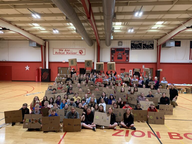 A large group of students in a gym holds up cardboard signs, some spelling "HOPE," with more displays in front of them—part of their effort to assemble Wheaton cancer care packages.