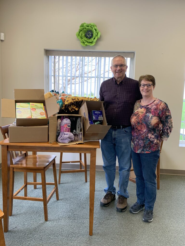 Two people stand beside a table with open boxes containing food and yarn in a well-lit room with a green decorative flower on the wall, assembling Orlando care packages for those in need.