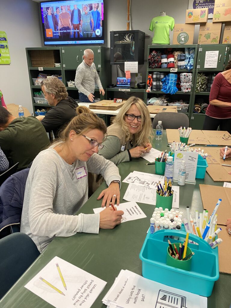 Several people sit at a table covered with papers and art supplies, writing and drawing cards for Orlando care packages. Others work and talk in the background of a classroom or workshop setting.