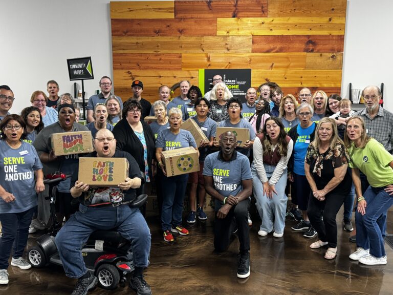 A diverse group of people poses indoors, many wearing “Serve One Another” shirts and holding decorated boxes with positive messages, highlighting Crown Point volunteer opportunities in front of wooden wall panels.