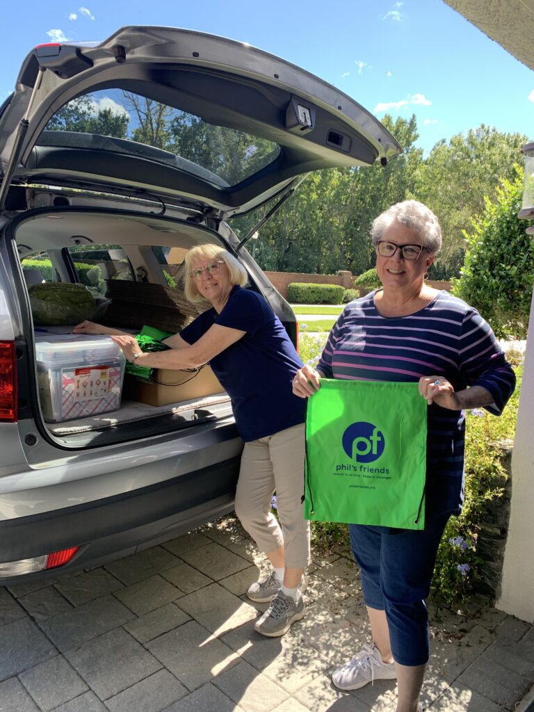 Two women stand by an open SUV trunk; one loads items while the other holds a green Phil’s Friends bag. It is a sunny day in a suburban neighborhood, highlighting Chicago faith-based cancer support in action.