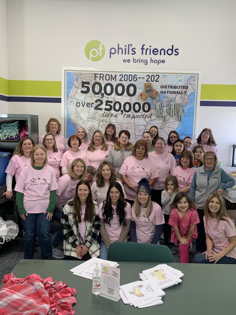 A group of women wearing pink shirts pose together in front of a Phil’s Friends sign and a large poster displaying statistics about lives impacted by Chicago faith-based cancer support and items distributed.