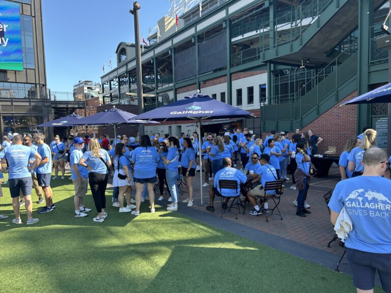 A large group of people in matching blue "Gallagher Gives Back" shirts gather outside near a stadium under umbrellas on a sunny day, coming together to support Crown Point volunteer opportunities.