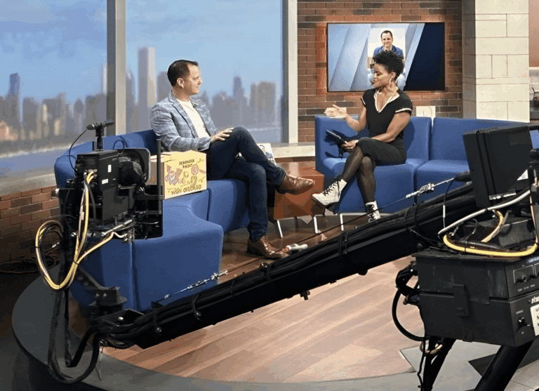 A man and a woman sit on blue couches in a TV studio, having a conversation about Chicago faith-based cancer support while cameras film the interview. A cityscape is visible through the window.