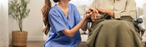 A healthcare worker kneels beside an elderly person in a wheelchair, holding their hands in a supportive gesture. The setting appears to be a home or care facility offering Chicago faith-based cancer support.