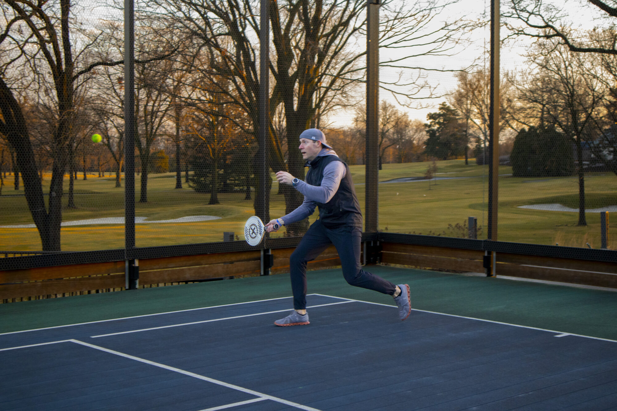 A person wearing athletic clothing plays paddle tennis on an outdoor court, preparing to hit a ball with a paddle. Trees and a golf course are visible in the background.