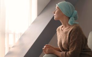 Woman with a headscarf sits indoors on a chair, hands clasped, looking thoughtfully toward a bright window.