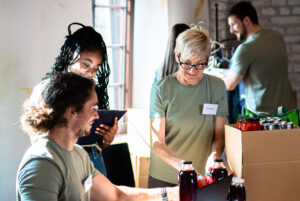 Three people packing food items into boxes, wearing name tags and casual clothes, in a bright room with shelves and canned goods in the background.