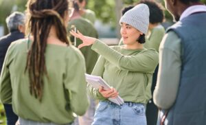 A person in a gray beanie and green shirt gestures while holding papers, talking to others outdoors in a group setting.