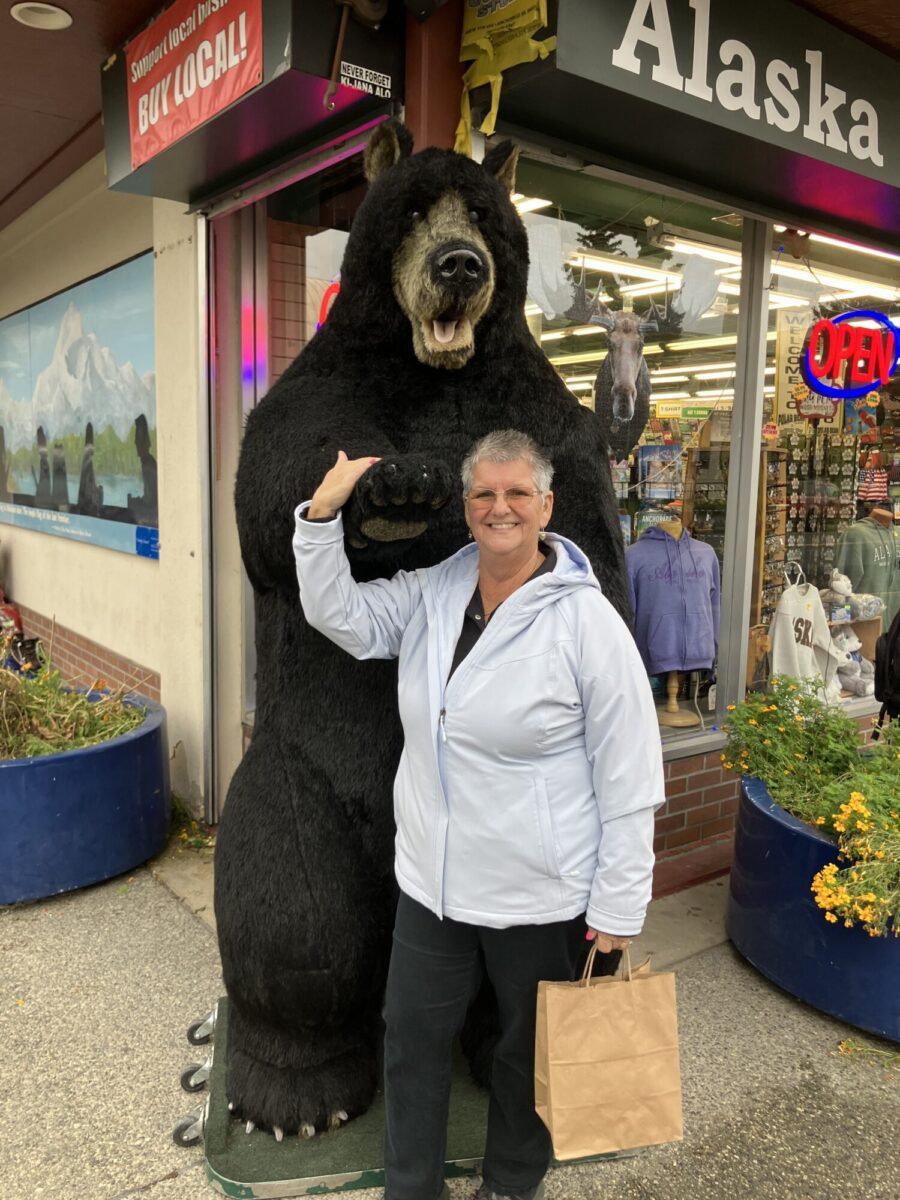 A person in a light jacket stands next to a large bear statue outside a souvenir shop in Alaska, holding a shopping bag and smiling at the camera.