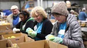 Four people sort and inspect produce in cardboard boxes at a food bank or distribution center, wearing gloves and outdoor clothing.