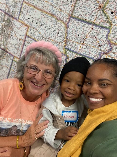 Three people smiling in front of a large map of Nebraska and Iowa; an older woman, a young boy with a name tag reading "Chase," and a younger woman wearing a yellow scarf.
