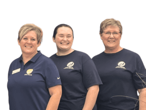 Three women wearing navy blue shirts with a logo stand side by side, smiling at the camera against a plain light background.