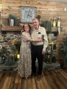 An older couple stands arm in arm in front of a stone fireplace with rustic decor, wooden walls, and a large clock above them.
