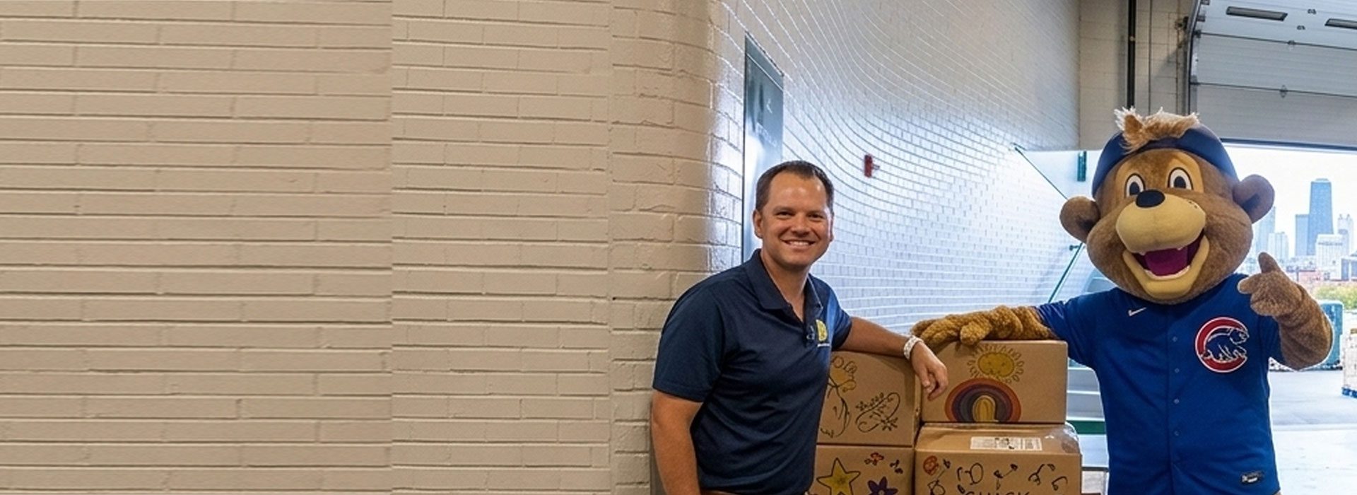 A man stands next to a person in a Chicago Cubs mascot costume, both beside decorated cardboard boxes in a warehouse loading area.
