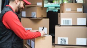 A delivery worker in a red cap and uniform signs a package among several cardboard boxes in the back of a van.