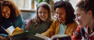 Four women sit together indoors, smiling and reading books, with natural light and plants in the background.